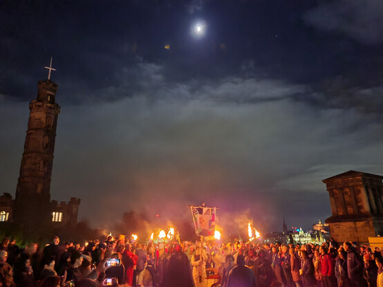 Part of the Beltane Fire Festival heads towards the camera, flaming torches and a banner held aloft the costumed and made up cast of fantastic characters are flanked by a crowd left and right, behind and above them a large moon lighting clouds in the sky, top left the Nelson monument tower sticks up and middle right an old stone building of the city observatory stares impassively down