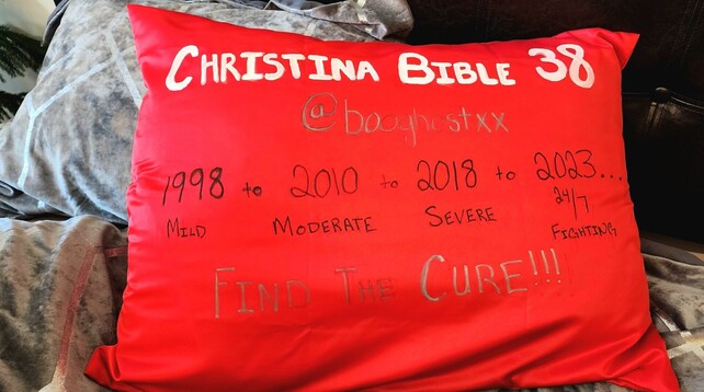 A red pillow case that says in large white letters “Christina Bible 38” and in smaller black letters “1998 mild to 2010 moderate to 2018 severe to 2023 24/7 fighting …” and in large silver letters at the bottom, “Find a cure!!!”