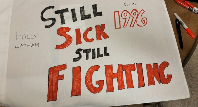 A white pillow case that says in red and black, Holly Latham Still Sick Still Fighting since 1996
