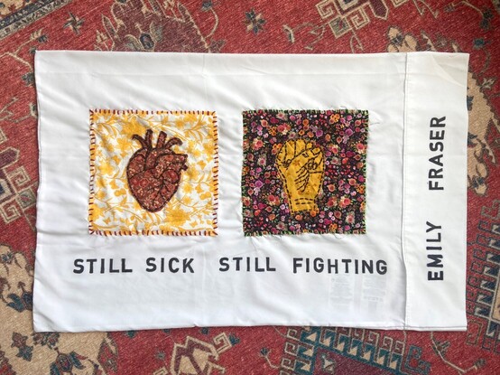
A white pillowcase with a yellow floral square of fabric on the left and a darker floral square of fabric on the right. There is a red anatomical heart sewn over the yellow square & a yellow fist sewn over the darker square. Below in black capital letters it says “Still Sick Still Fighting.” “Emily Fraser” is written across the right edge of the pillowcase.