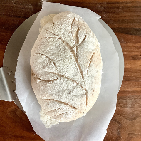 An unbaked bread with a design carved into it, on baking paper on top of dark wood.