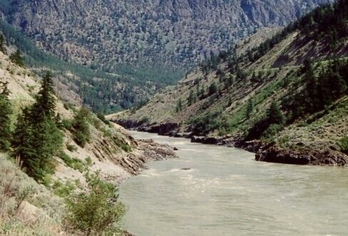 Rapid filled river flows between rock and steep banks toward the high, semi-tree covered hills in the background. 