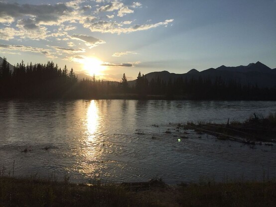 Sunrise or sunset over a smooth body of water. In the mid distance a line of hills and trees are silhouetted against a lighter sky.