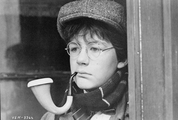 A still frame from the film “Young Sherlock Holmes” featuring actor Alan Cox who portrayed John Watson, looking off frame as he smokes a pipe. 