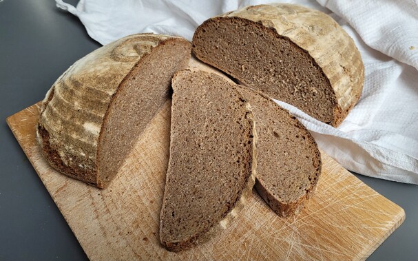 Auf einem Schneidebrett liegt das Brot vom letzten Post teilweise aufgeschnitten, eine Hälfte liegt neben dem Brett auf einem weißen Leinen Tuch. Zwei Scheiben liegen in der Mitte des Bildes, man sieht die gleichmäßige Porung. 