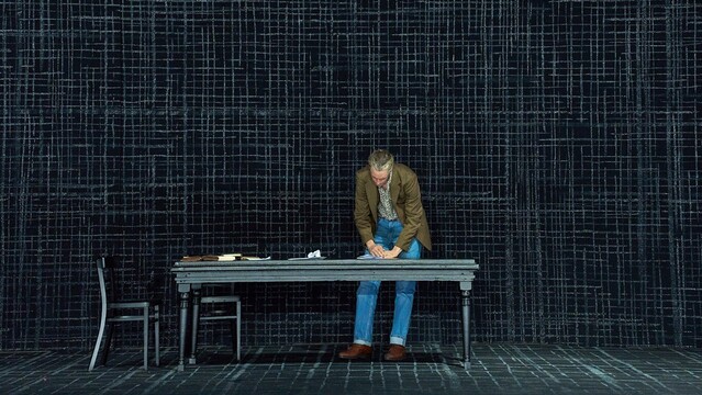 Dark stage with black walls, on them a grey cross-hatch pattern reminiscent of coarse fabric. Black dining table with black chairs, books and crumpled letters on it. Behind the table stands a tall man with blond hair wearing a brown jacket, blue jeans, a patterned shirt and brown boots, hunched over, looking at a book on the table. Baritone David Pichlmaier as Eugene Onegin in Isabel Ostermann’s 2023 staging of Tchaikovsky’s opera Евгений Онегин at the Staatstheater Darmstadt. Photo © Nils Heck / Staatstheater Darmstadt