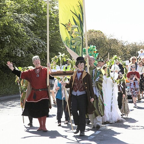 A parade through an English village, people in fancy dress including mediaeval robes, fairy costumes, and top hats, carrying flower garlands and an embroidered banner depicting cowslips