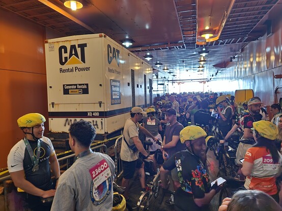 The automobile deck of a ferry is packed with bicyclists. a large generator trailer sits in a fenced off area to the side 