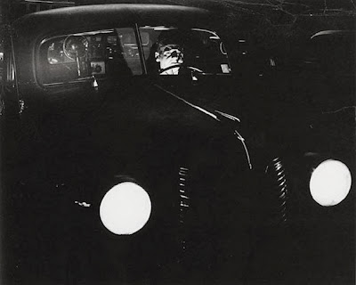 ominous black and white photo of a motorist in a vintage car at night, headlights on.