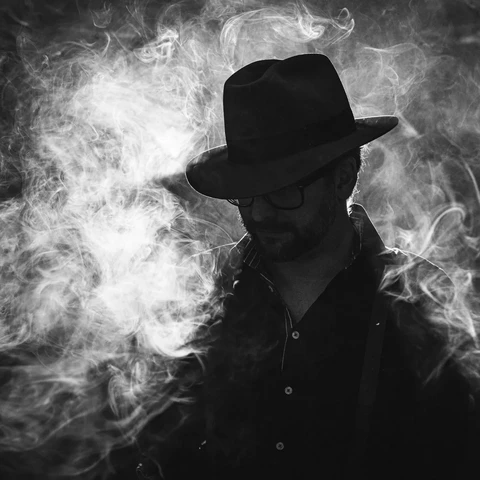 Black and white photo. Portrait of a man taken in a photo studio, upper body. Man's face is hidden by cowboy hat. Noire vibes. Smoke is illuminated and the man is mainly visible as a silhouette.