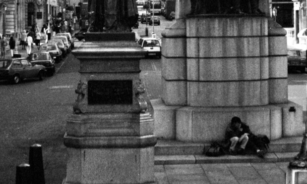 A monochrome street photo of a couple snuggling at the base of mid-street statues in Waterloo Place, London while traffic and pedestrians carry on around them