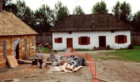Two post and sill buildings in a fur trade fort. One is whitewashed, both are shingle-roofed. These buildings are in the replica Fort Langley. 