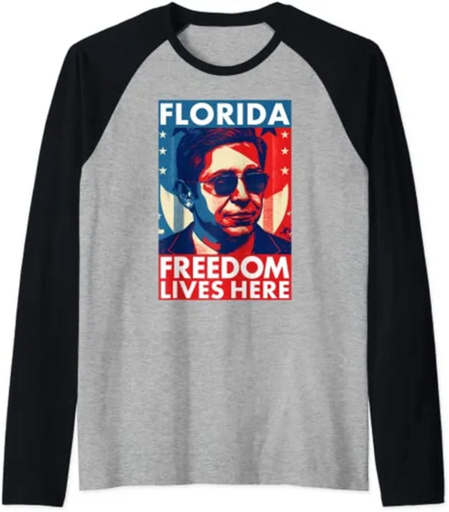 Ein Sweatshirt in grau und schwarzen Ärmeln. Auf dem Vorderteil in Grau ist ein Foto von Ron DeStantis und der Aufdruck „Florida Freedon Lives here“. Der Aufdruck und das Gesichts von DeSantis sind in rot weiß blau.