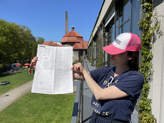 Person with hat with the letters A MAZE. holding a paper with a schedule and a map. Field of grass and buildings in the background.