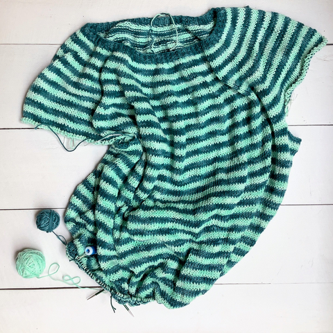 Flat lay view of a striped circular yoke garment in teal and light turquoise, shown on a circular knitting needle on a white plank background together with the 2 balls of yarn.