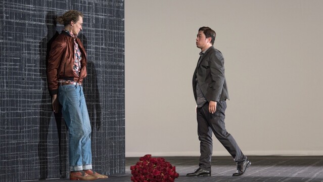 Two men on a stage. On the left a tall blonde man with his long hair in a bun, wearing a bronze-coloured silk windbreaker, a shirt with a colourful print, blue jeans and two-tone leather shoes, is pressed against a black wall with a light grey crosshatched pattern reminiscent of coarse fabric. In the center a dark-haired man in a grey suit and black patent leather shoes approaches him cautiously from the brightly lit open stage. Between them on the floor a large bouquet of red roses.
Baritone David Pichlmaier as Eugene Onegin and Tenor David Lee as Vladimir Lensky in Isabel Ostermann’s 2023 staging of Tchaikovsky’s opera Евгений Онегин at the Staatstheater Darmstadt. Photo © Robert Schittko / Staatstheater Darmstadt