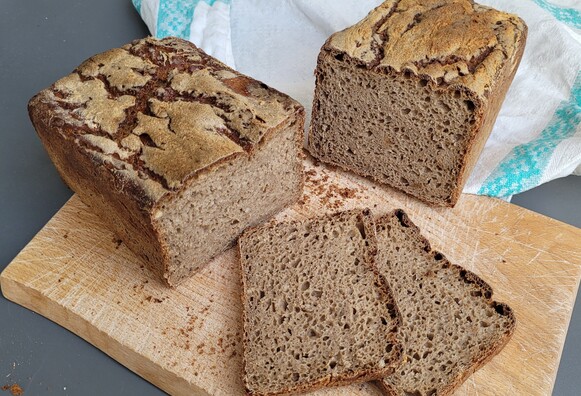 Ein Kastenbrot liegt auf einem Schneidebrett, in der Mitte geteilt, zwei Scheiben sind abgeschnitten. Die Krume ist mittelporig und einigermaßen gleichmäßig. Die Kruste ist oben leicht bemehlt und unregelmäßig eingerissen. Im Hintergrund liegt ein Leinentuch. 