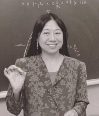 Black and white photo of Sau Lan Wu standing in front of a chalkboard. She is wearing a floral jacker, and is holding her right hand up at roughly shoulder height with her index finger touched to her thumb. She has long black hair, is smiling, and is looking at the camera. Black and white photo of Sau Lan Wu standing in front of a chalkboard. She is wearing a floral jacker, and is holding her right hand up at roughly shoulder height with her index finger touched to her thumb. She has long black hair, is smiling, and is looking at the camera.