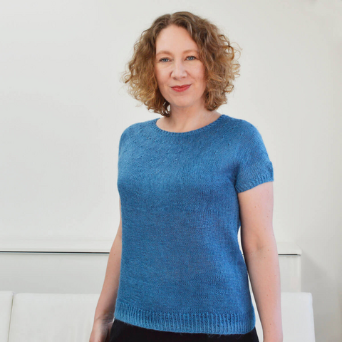 A woman with short curly hair, wearing a blue handling fine-gauge tee, and standing in front of a white couch and wall.