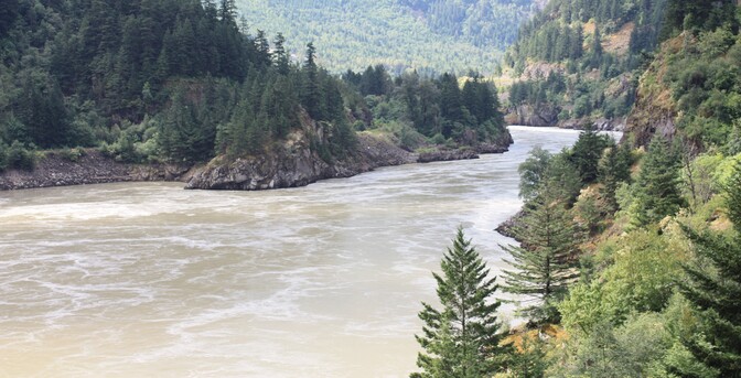 A whirlpool filled river flows around a sharp bend in the rocky river toward the photographer. The river banks are heavily treed, and the hills behind are also well forested.
