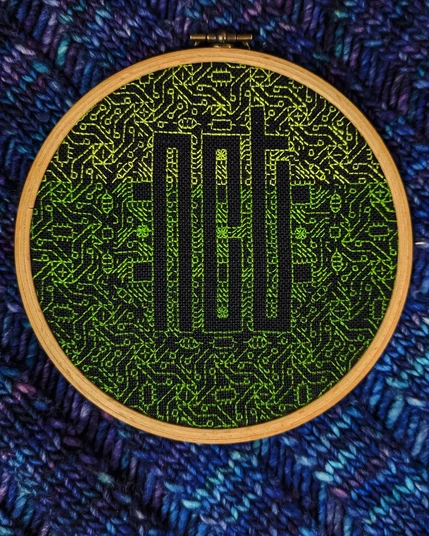 A gradient green embroidered background framing the NCT logo on black linen evenweave in a small hoop.