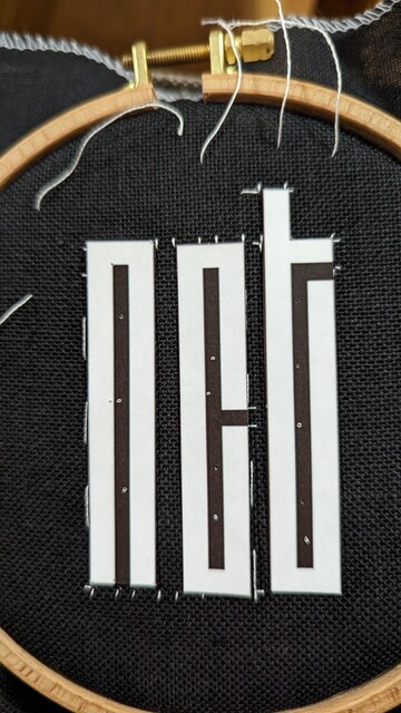 White thread crafting an outline of the NCT logo on the black linen cloth. The NCT letters cut out from paper.