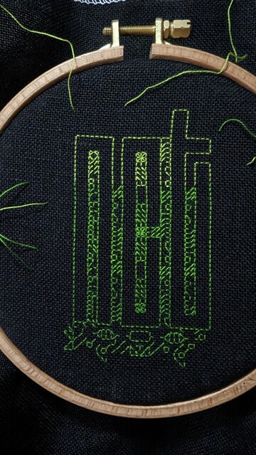 The NCT logo is outlined in embroidery now and the spaces between slowly filling up with pattern against black linen evenweave.