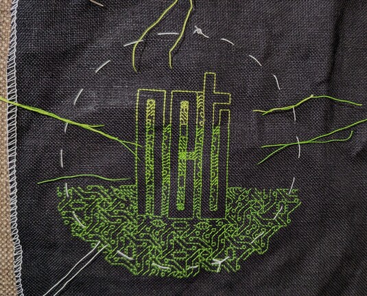 A green gradient NCT logo embroidered with circuitry embroidery on black linen. White outline is the shape of the hoop, and stitching slightly outside of it so that I have embroidered leeway when I finish the hoop.