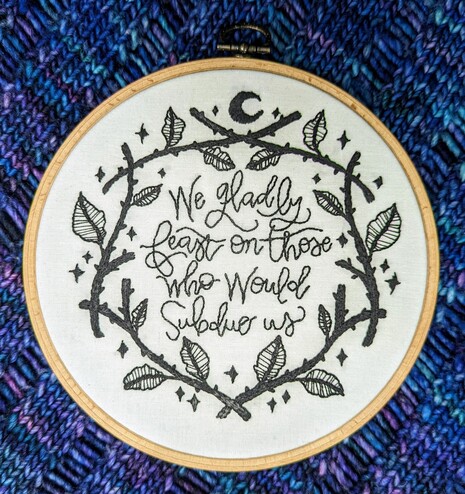 Black embroidery on white cotton cloth with the quote "We gladly feast on those who would subdue us" surrounded by a 'wreath' of thorns and leaves and stars. It is finished in a hanging round frame.