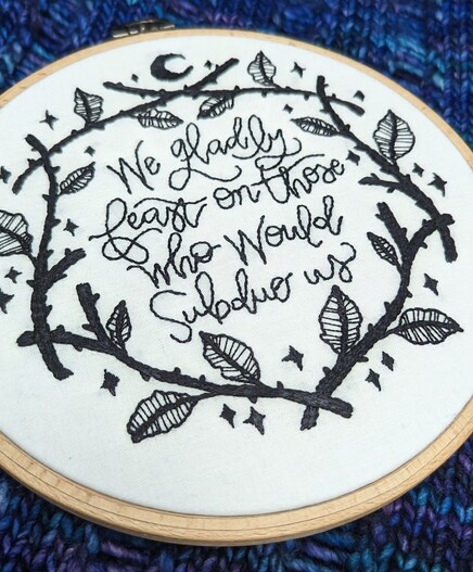 A closer look at black embroidery on white cloth that reads "We gladly feast on those who would subdue us" survived by a wreath of thorns and leaves and stars. 