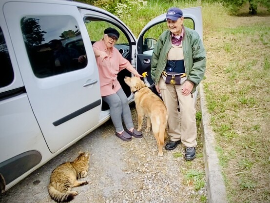 Elderly couple and animals next to car. Big fluffy tabby cat laying down next to the rear wheel. My mother seated on the passenger side with her feet outside pointing at me so the dog at her feet would look at the lens. My dad stands next to them and smiles before the open side door. 