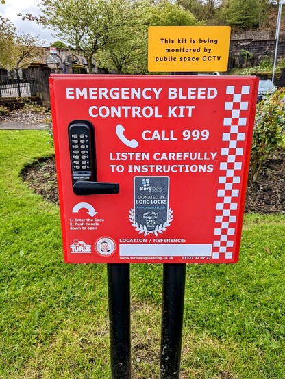 The red sign is attached to 2 black poles in the ground. The black object on the left of the red board is a keypad and below that is a black handle. The person who calls 999 is given the pin number and the box can then be opened to retrieve the kit inside.
Behind the red box to the left is the arches of the railway station, you can just make out the old British Rail logo.