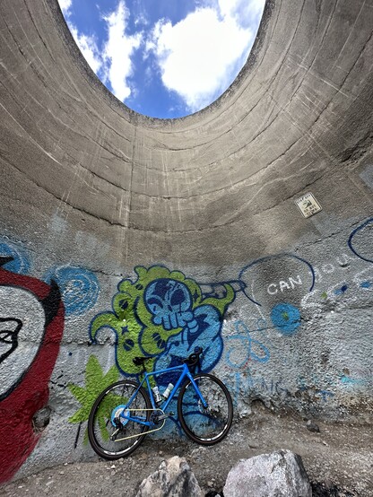 A blue gravel bike against a graffiti wall with blue skies and clouds visible above