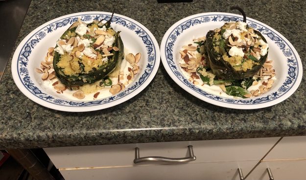 Plated dish. Roasted poblano pepper stuffed with couscous, spinach, golden raisins, roasted red pepper, feta, and tahini dressing sitting on top of lemon yogurt. Garnished with more feta and sliced toasted almonds.