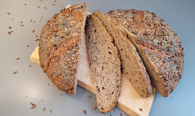 Das Brot liegt, in der Mitte durchgeschnitten und mit zwei abgeschnittenen Scheiben, auf dem Schneidebrett. Man sieht die etwas ungleichmäßige, mittelporige Krume und die Leinsamenkörner darin. 