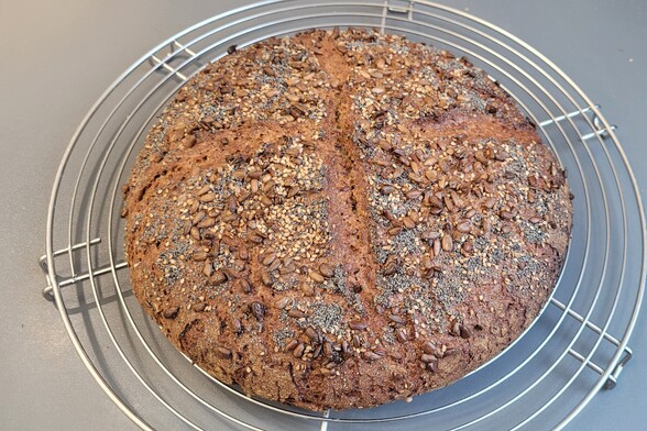 Ein runder Laib Brot auf einem Kuchengitter, die Kruste ist vollkorntypisch, nicht zu dunkel, kreuzförmig eingeschnitten und außerhalb der Schnitte mit dem beschriebenen Saatenmix bedeckt. 