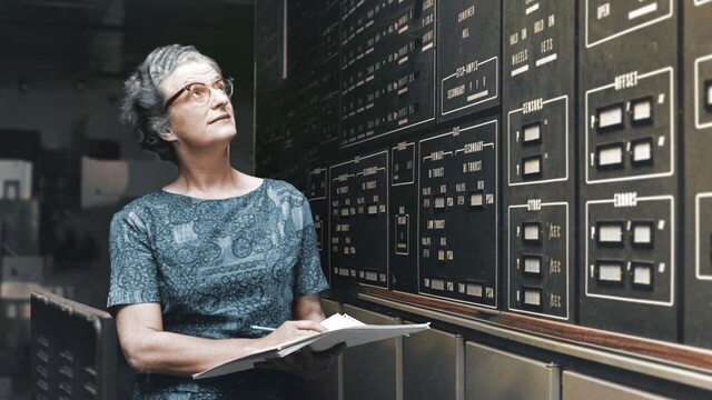 A color photo of Nancy Grace Roman. She is standing next to a bank of analog instruments, which she is looking at while writing on a stack of papers held in her left hand. She has on a grey-blue top with an intricate design, and is wearing glasses. Her short, grey hair is wavy. The bank of instruments features several needle readouts, clustered in groups of two, three, or four.
