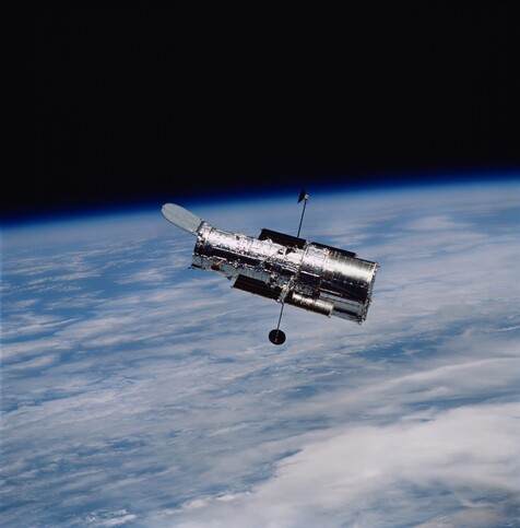 A color photo of the Hubble Space Telescope, taken by the crew of one of the shuttle missions. The Hubble is a mostly silver cylindrical tube, with the bright blue and white curve of the Earth below. One end has a raised flap, and two thin arms sporting small dishes extend from the sides.