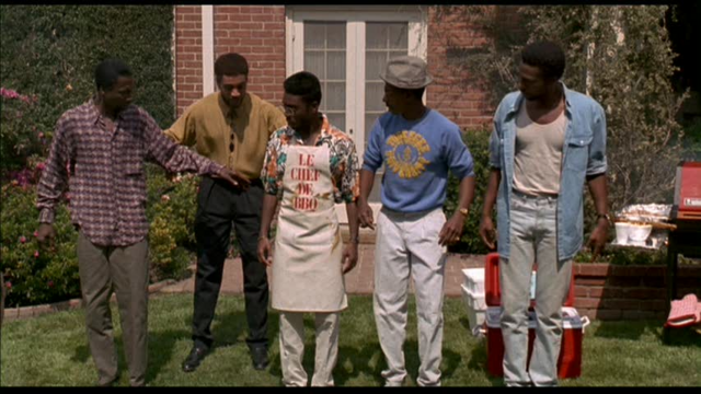 Five African-American men in their 40s are attending a backyard barbeque. Behind them is a brick house. To their right is a barbeque grill. They are preparing to do a dance number they did when they were younger and members of a famous Motown-style R&B group called The Five Heartbeats, and the three on the right are looking at the two others for direction. The man in the middle is wearing an apron that reads "Le Chef de BBQ".