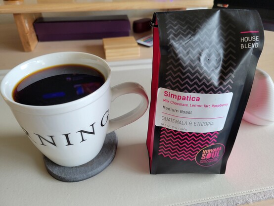 16 fluid ounce white mug of black coffee on a gray coaster on a white desk mat, to the left of a 12 ounce pink and black pouch of coffee beans. The mug partially shows text Good Morning. The coffee pouch reads "Simpatica / Milk Chocolate, Lemon Tart, Raspberry Medium Roast / Guatemala & Ethiopia / Net weight 12 oz 340 g / Nirvana Soul Coffee Purveyors"