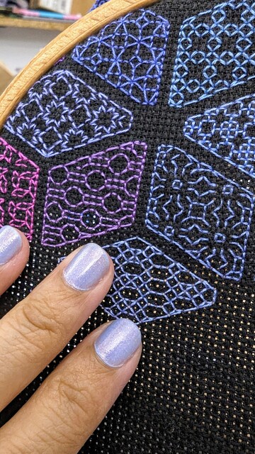 In progress blue and purple and pink blackwork embroidery diamonds on black Aida cloth in a hoop. Two fingers with glittery nail polish match one of the diamonds.