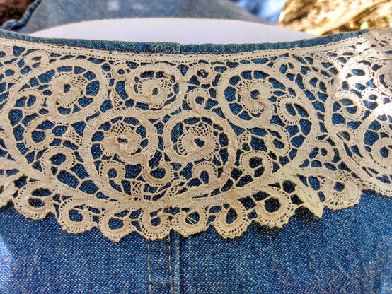 A close up look at the antique Cantu lace collar. It has swirly vines that wind up in a small sort of 3-petal flower motif where the end of a swirl ends up. The gentle curvy vines connect to adjacent ones with tiny braid connections. The linen is a beige almost tea-dyed hue, and it contrasts with the washed blue denim of the vintage jacket.