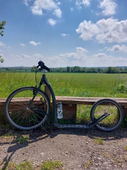 A grey kickbike leaning against a rustic wooden bench in front long grasses and rolling fields. There are trees in the background, and the sky is a pale blue and scattered with clouds.