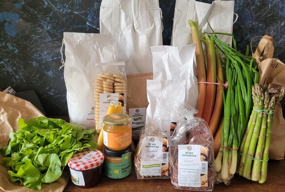 Die Einkäufe der Tour auf einer Holzfläche aufgebaut: versch. Mehlsorten, zwei Sorten Getreide, Kartoffeln, Eier, Honig, Orangensenf, Konfitüre, Kekse, Bienenwachskerzen, ein Salat, Rhabarber, Frühlingszwiebelm, grüner Spargel. 