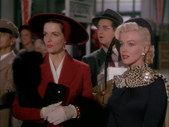 Screenshot from 1953 film “Gentlemen Prefer Blondes” with Jane Russell in a red jacket, white gloves and mink scarf, and Marilyn Monroe in black dress and leopard skin scarf. 