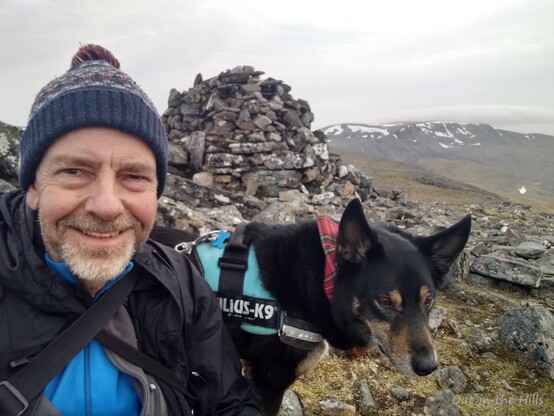 Selfie of myself and Moray the dog sat by a summit cairn. More hills in the background with some snow patches. 