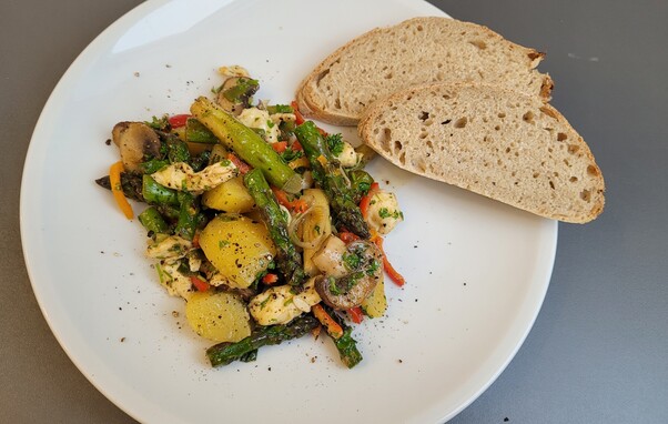 Foto: Der Salat auf einem weißen Teller. Außer den genannten Zutaten sind noch Kartoffelstückchen, rote und orangene Paprika Streifen und Petersilie darin. Darüber ist Pfeffer gestreut und auf dem Teller liegen noch zwei Scheiben des Weißbrots.
