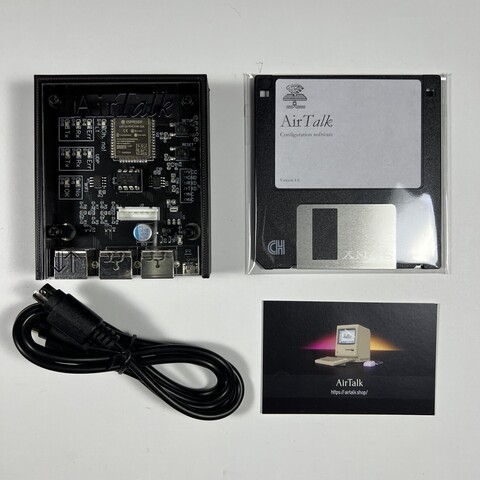 An AirTalk module next to a floppy disk, cable, and business card. The module itself is a small black case with a clear top showing a circuit board inside with a label that says AirTalk on top. 