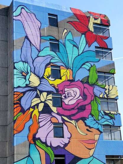 Street art in vibrant red, purple, pink, yellow & green on the front of an ugly 20th century office building. A woman's head decorated with flowers.