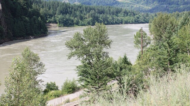 A whirlpool filled river flows between rocky, tree-lined banks toward the photographer. In the distance, more high hills covered with trees.